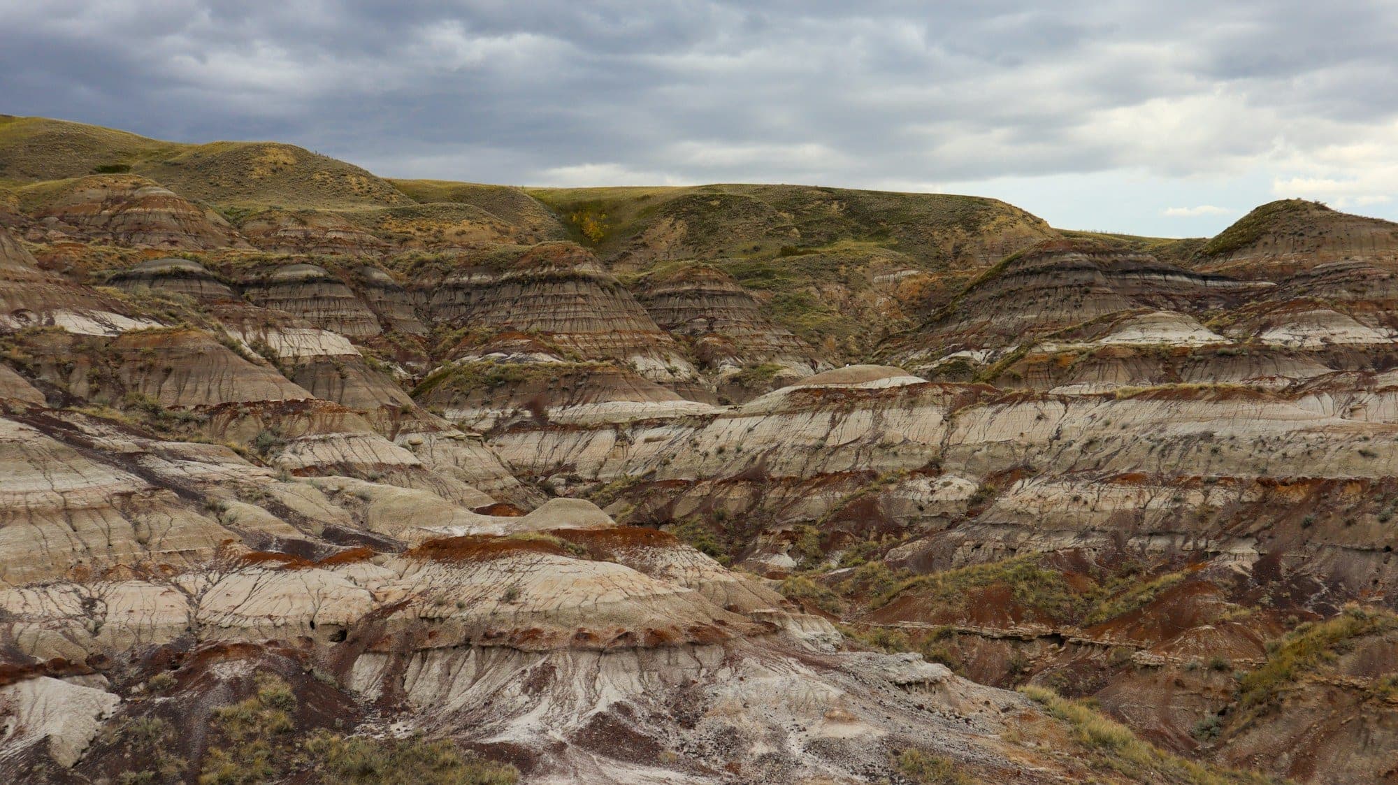 Layered red, grey and cream bands of the Drumheller badlands under a cloudy sky